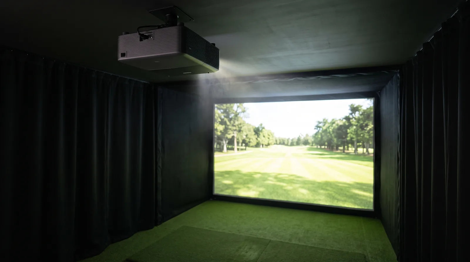 A ceiling-mounted projector throwing its beam onto the impact screen of a dark home golf simulator bay, with a fairway on the screen